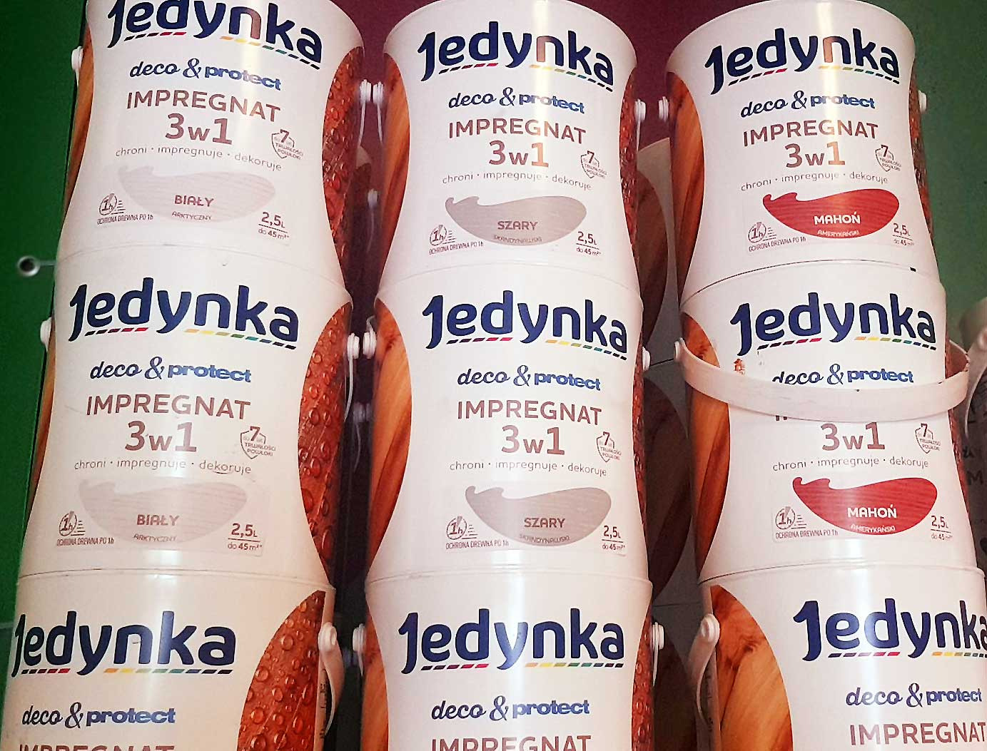 Półka w sklepie z farbami, na której stoją ustawione jedna na drugiej puszki z impregnatem do drewna firmy Jedynka w trzech odcieniach: białym, szarym i mahoniowym.