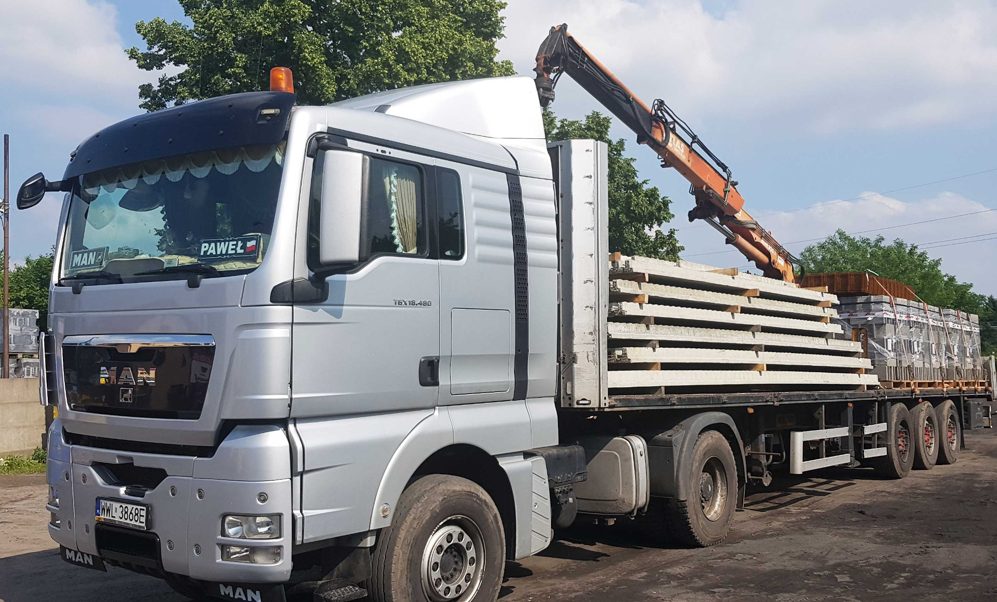 Ciężarówka MAN z dźwigiem przewozi palety z prefabrykatami betonowymi na platformie, widok z boku na tle zieleni i błękitnego nieba.