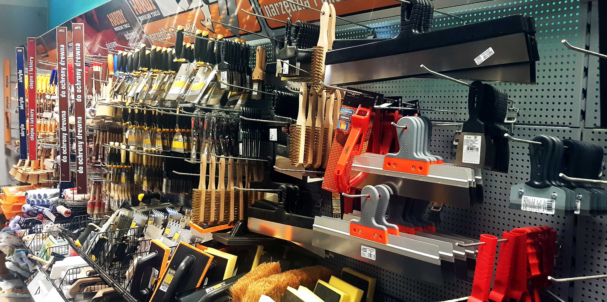 Ekspozycja różnorodnych narzędzi budowlanych i malarskich w sklepie: pędzle, szpachle, szczotki druciane, wałki i akcesoria do ochrony drewna, uporządkowane na metalowych regałach z perforacją.
