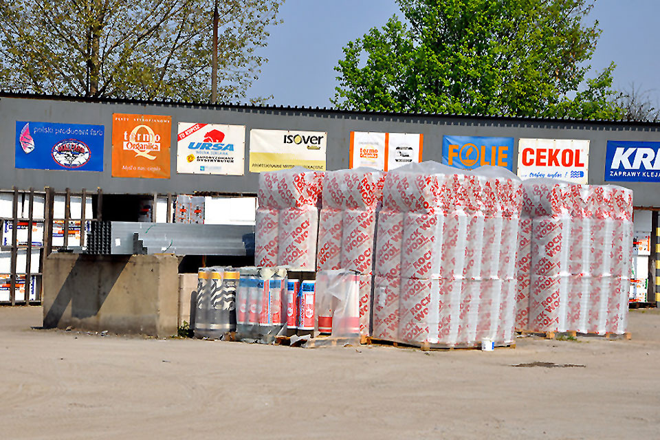 Stosy materiałów izolacyjnych Rockwool na paletach przed budynkiem składu budowlanego z reklamami producentów farb, systemów ociepleń i zapraw klejących.