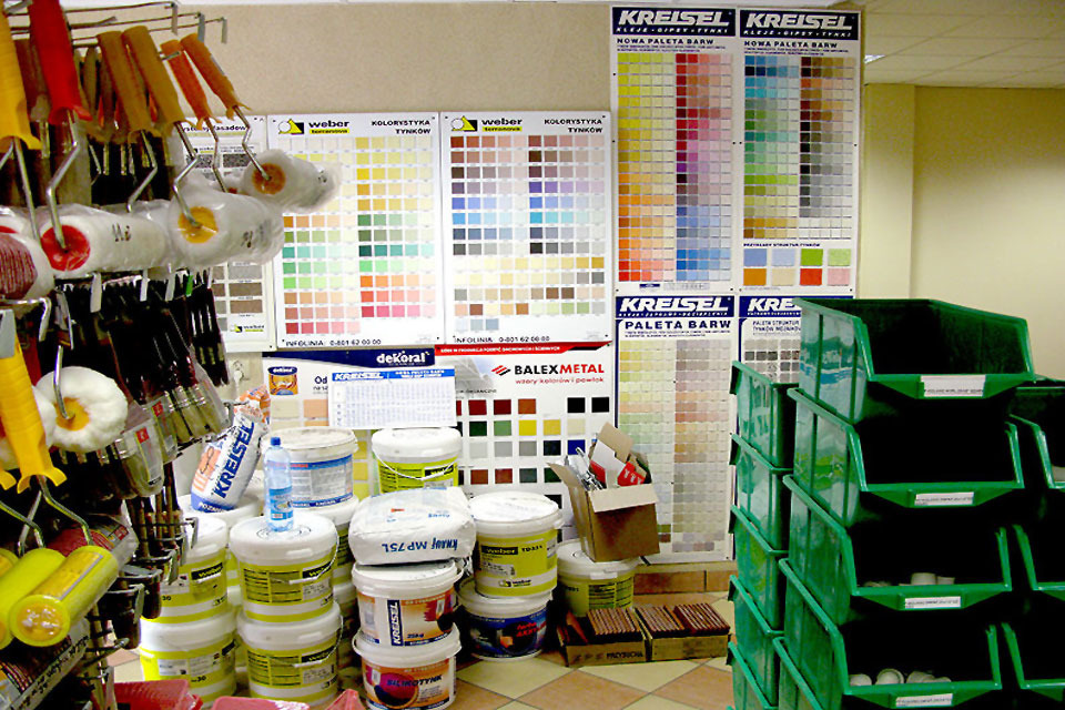 Wnętrze sklepu budowlanego: pędzle, wałki malarskie, farby w wiadrach, wzorniki kolorów różnych producentów oraz zielone pojemniki na drobne akcesoria malarskie.