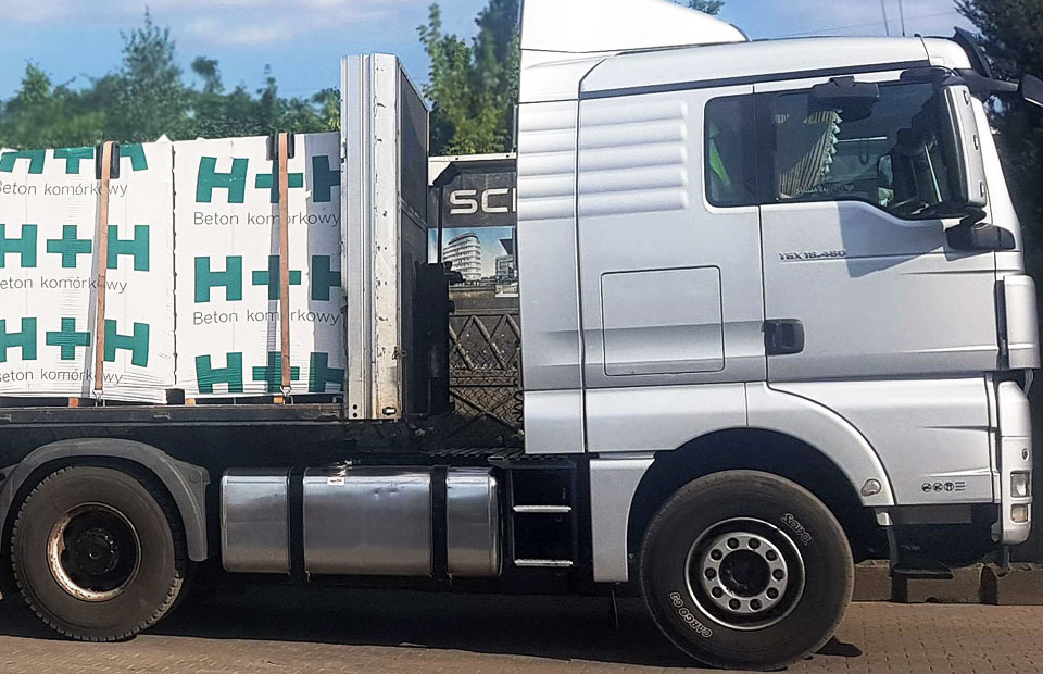 Ciężarówka MAN z załadowanymi paletami bloczków z betonu komórkowego marki H+H, zabezpieczonymi pasami transportowymi.