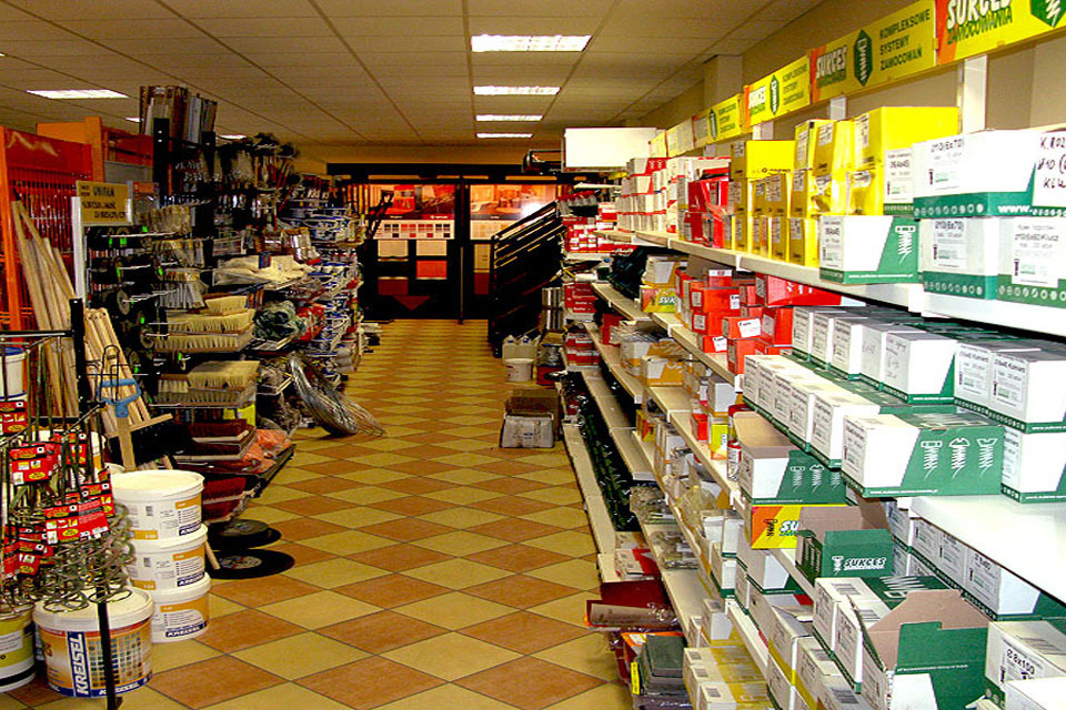 Wnętrze sklepu budowlanego z rzędami półek wypełnionych farbami, pędzlami, narzędziami i materiałami budowlanymi. Na pierwszym planie widoczne białe wiadra z farbą marki Kreisel.