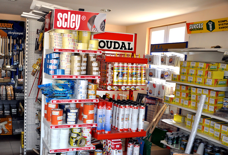 Wnętrze sklepu budowlanego z regałami pełnymi taśm malarskich, klejów, uszczelniaczy i innych materiałów wykończeniowych, z logo marek Scley i Oudal na górnych półkach, widoczne narzędzia malarskie...