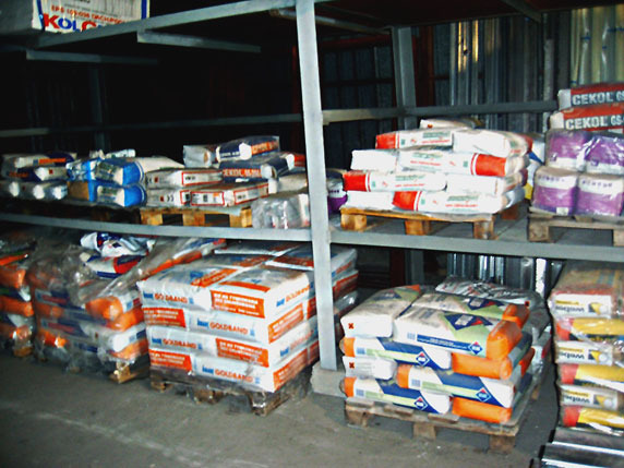 Regał magazynowy wypełniony paletami z workami zapraw budowlanych różnych producentów, widoczne etykiety Cekol, Goldband, ułożone warstwami na drewnianych paletach.