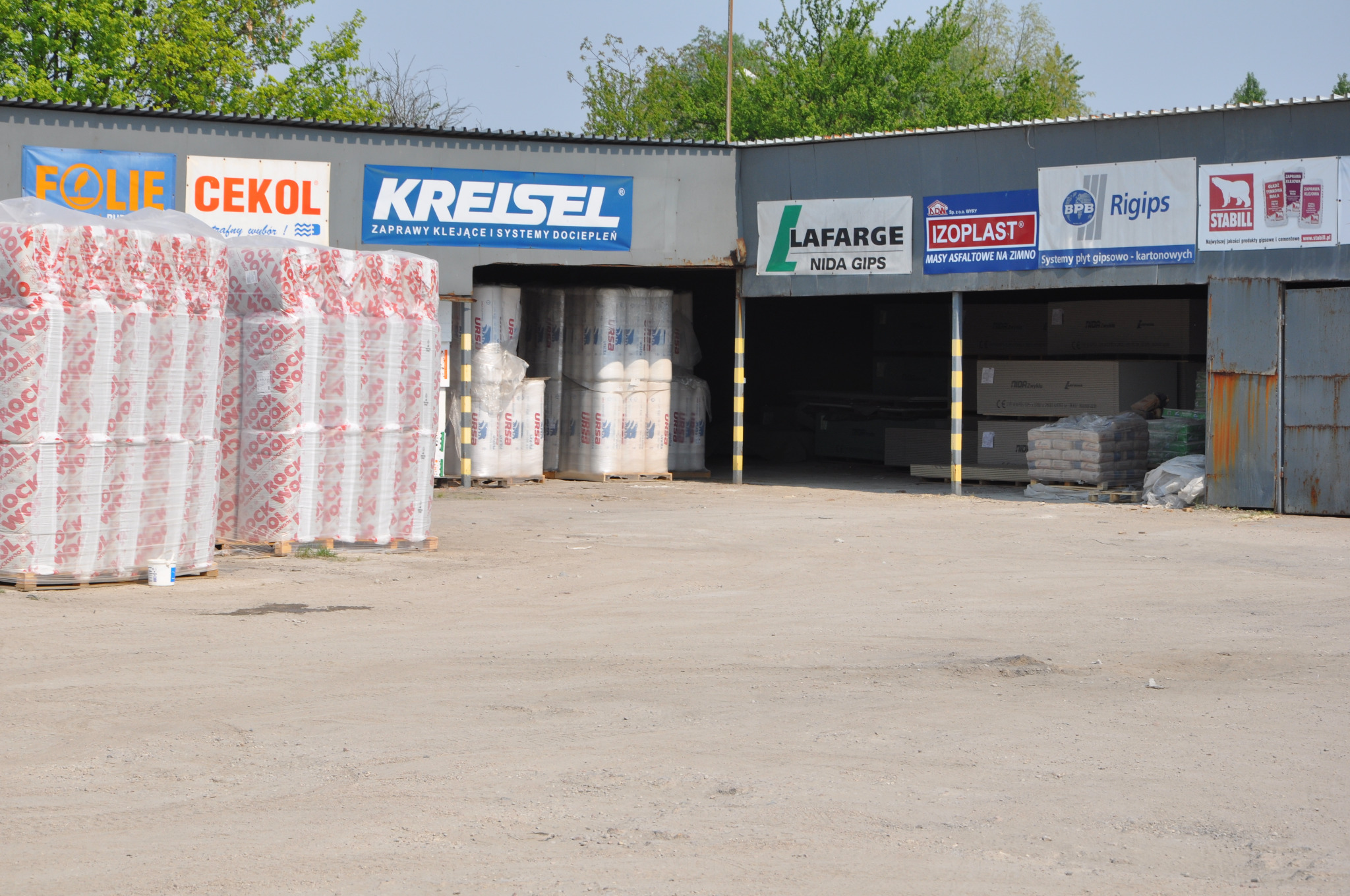 Skład budowlany na zewnątrz: palety z materiałami izolacyjnymi, widoczne logo producentów na budynkach magazynowych.