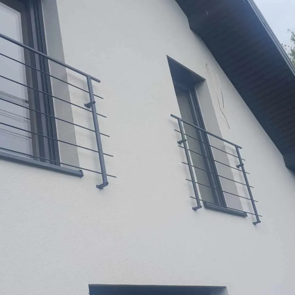 Minimalistyczne, czarne balustrady okienne z metalowych prętów, zamontowane na białej fasadzie budynku w Warszawie.