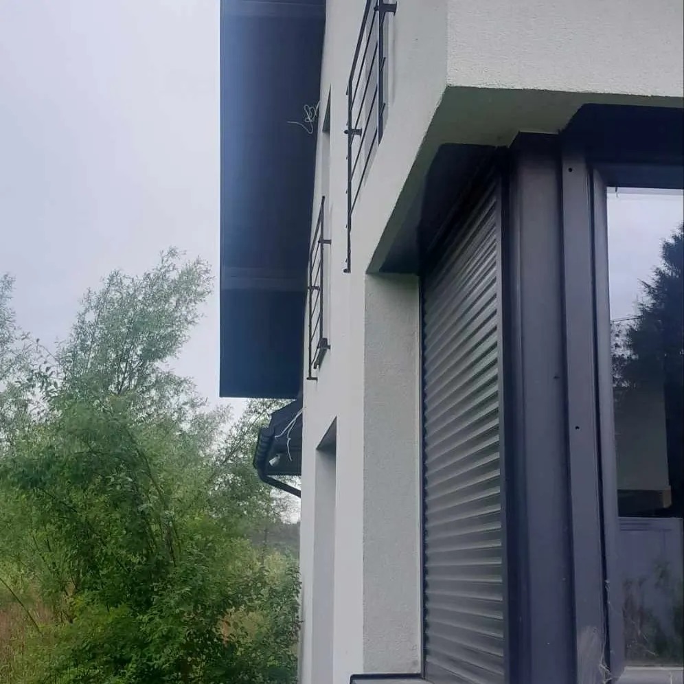 Fragment elewacji domu z czarnymi balustradami balkonowymi i roletami zewnętrznymi, widok z dołu, na tle zieleni.