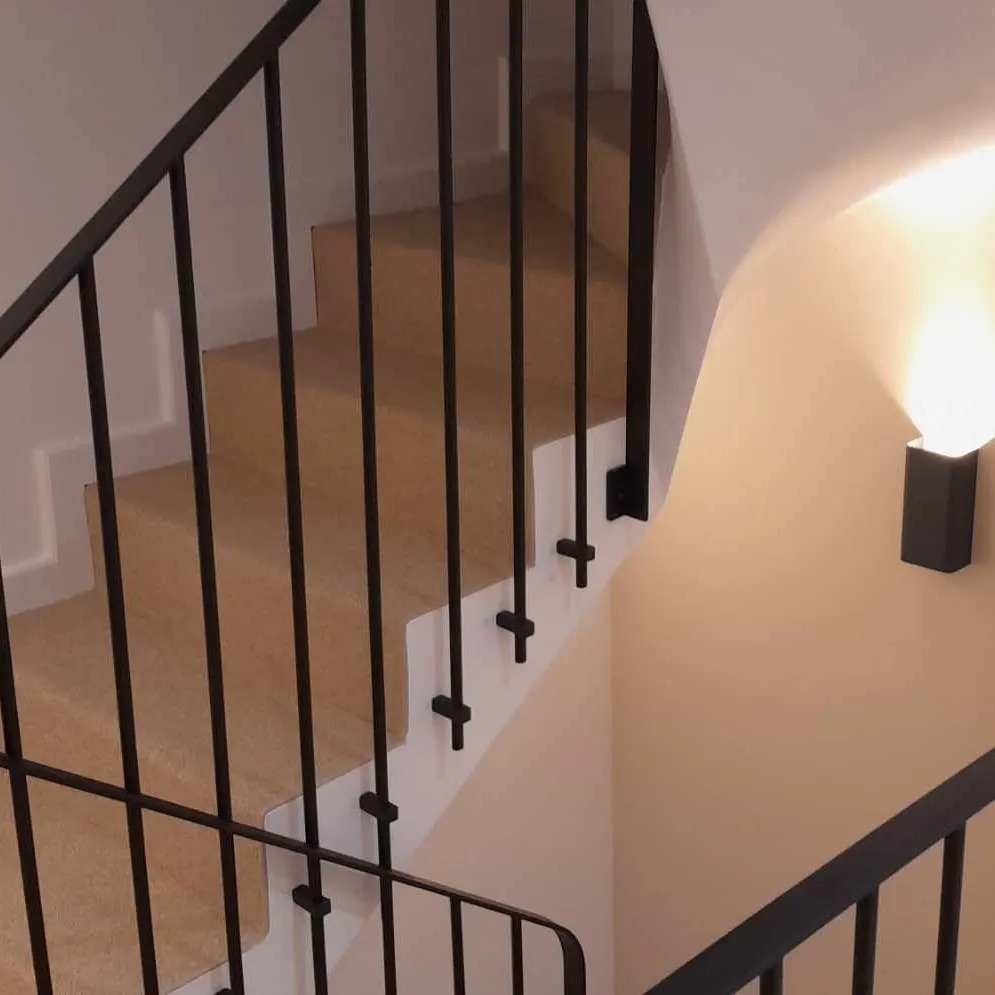 Nowoczesne schody wewnętrzne z drewnianymi stopniami i czarną, minimalistyczną balustradą z metalu, oświetlone kinkietem ściennym.
