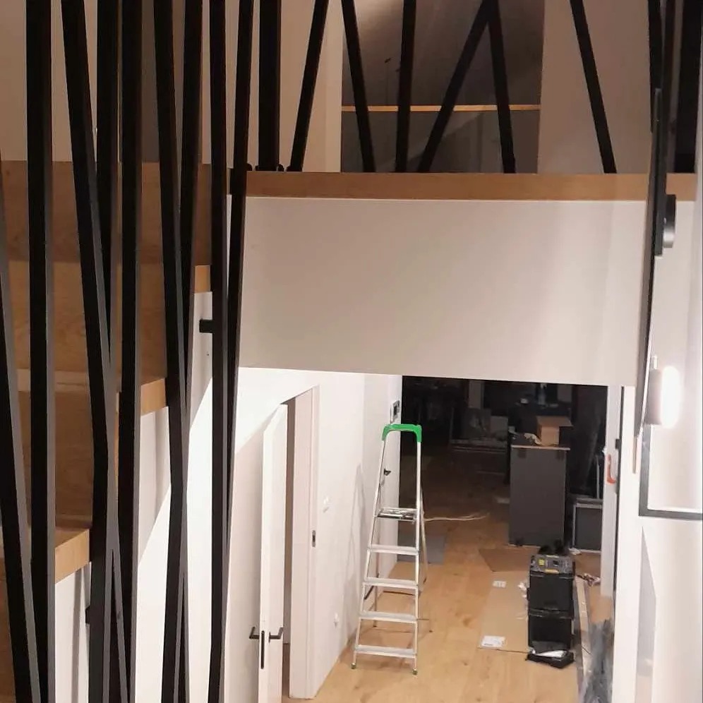 Nowoczesne schody z drewnianymi stopniami i czarną, pionową balustradą, widok z góry na hol z drabiną i częściowym bałaganem podczas remontu.