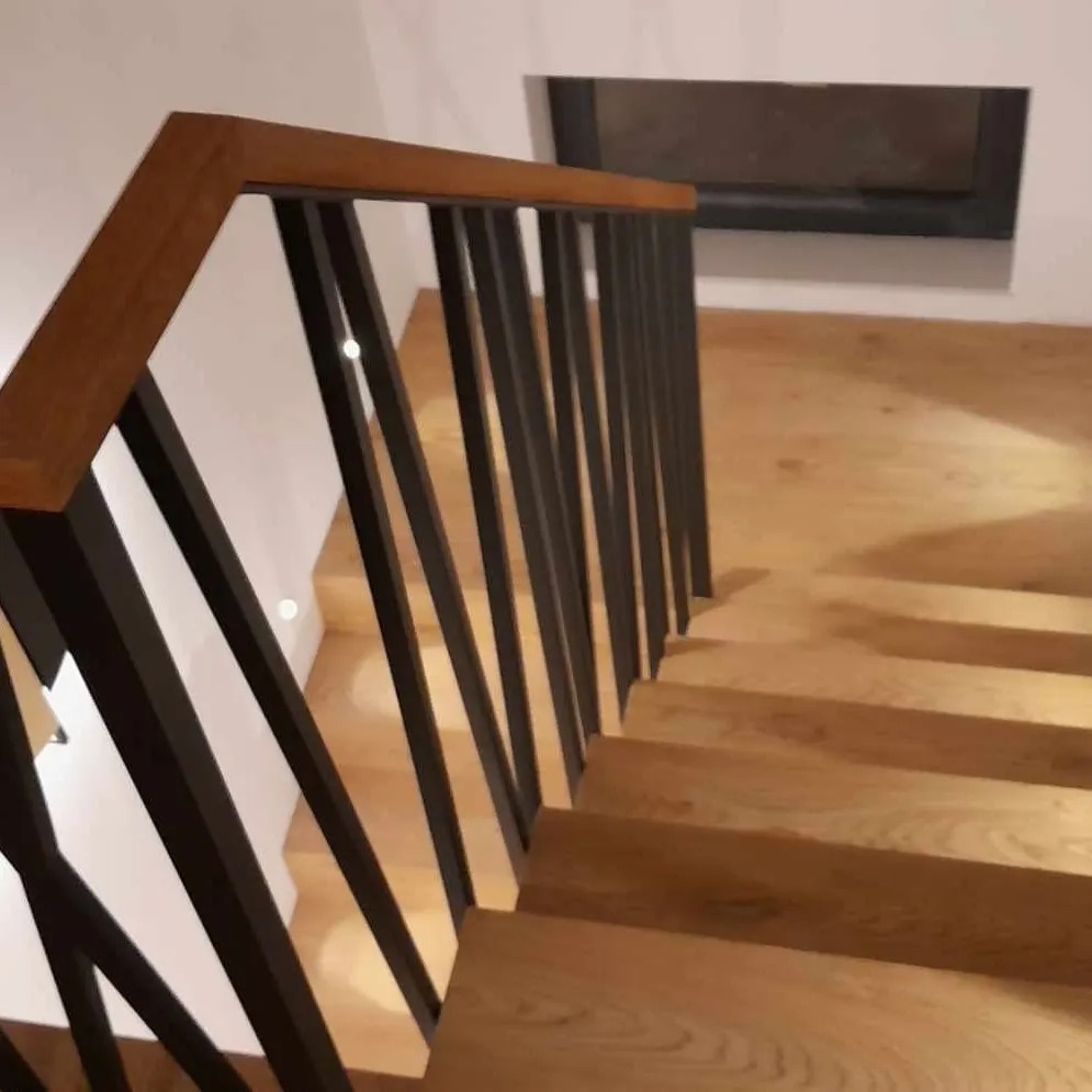 Nowoczesne drewniane schody z czarną, pionową balustradą i drewnianą poręczą, oświetlone punktowymi lampami w ścianie.