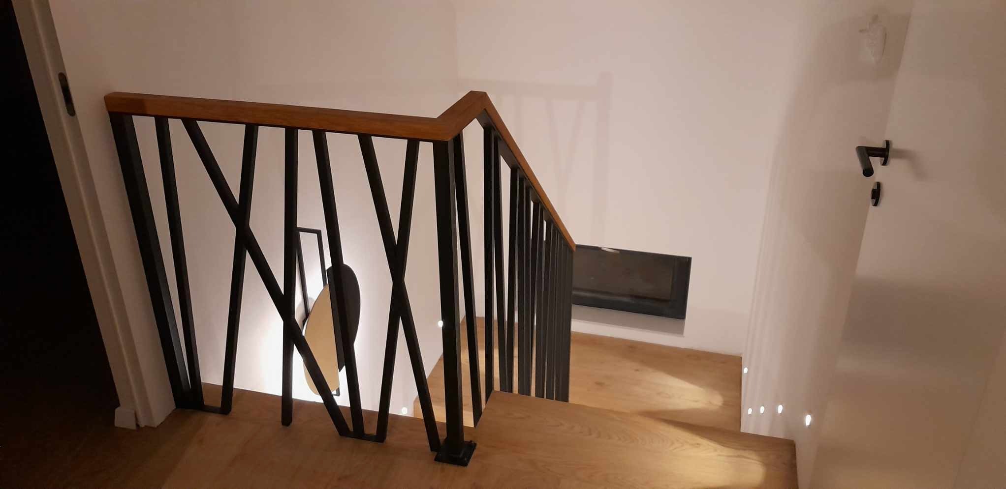 Nowoczesne schody z czarną, geometryczną balustradą i drewnianym poręczem, widok z góry na schody z oświetleniem punktowym w podłodze.