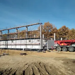Budowa domu z białych bloczków, widoczne rusztowania i żuraw samochodowy na tle jesiennych drzew i błękitnego nieba.