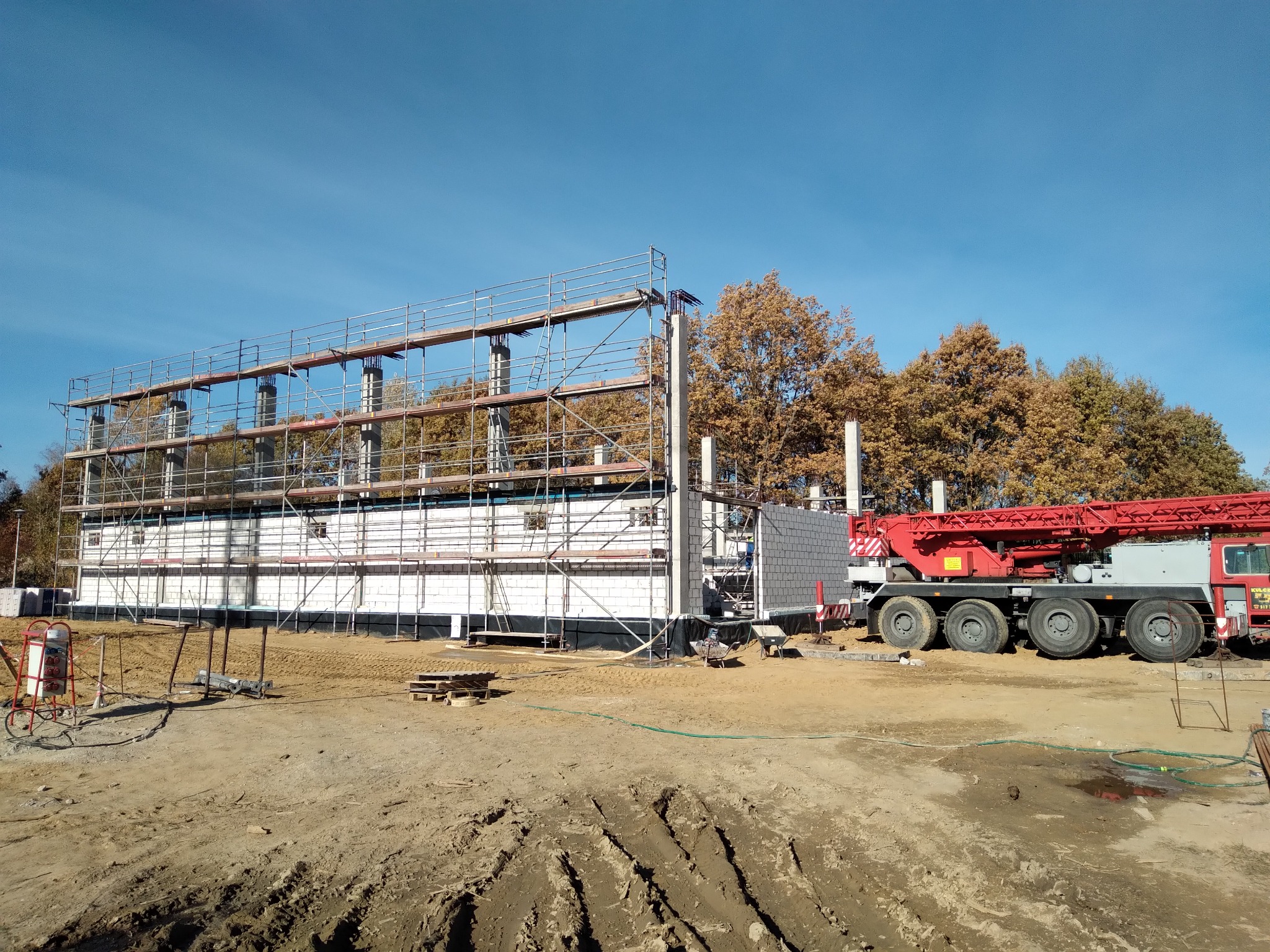 Budowa domu z białych bloczków, widoczne rusztowania i żuraw samochodowy na tle jesiennych drzew i błękitnego nieba.