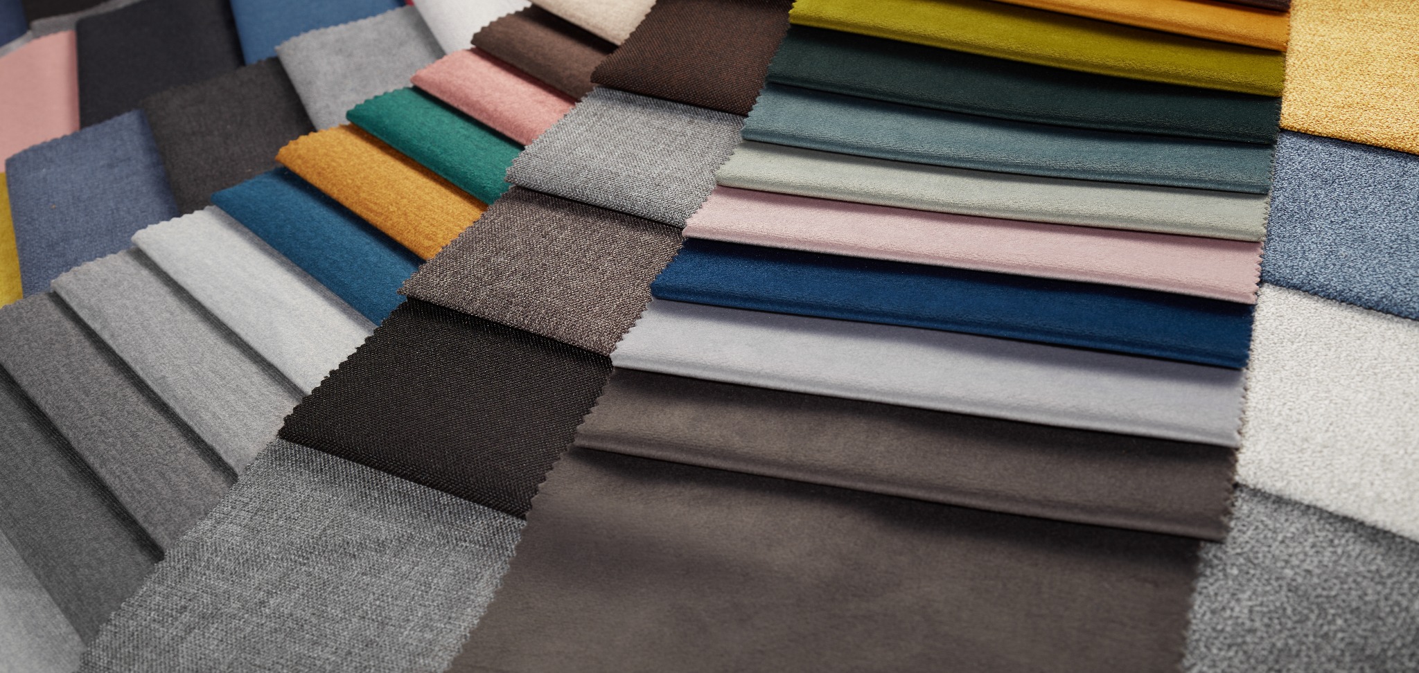 Próbki różnorodnych tkanin ułożone warstwami, prezentujące bogatą paletę kolorów i faktur, od szarości i brązów po błękity i róże, z delikatnym kontrastem pomiędzy tkaninami gładkimi a tymi...