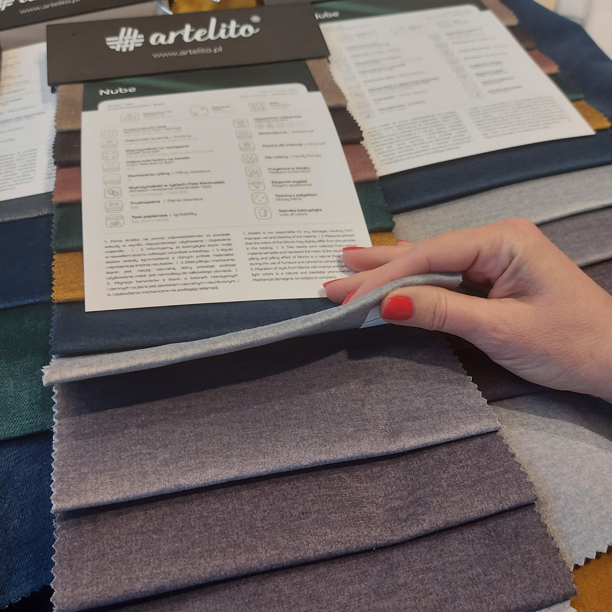 Próbki tkanin w odcieniach szarości i zieleni, prezentowane obok karty z informacjami o właściwościach, trzymane w dłoni z czerwonymi paznokciami.