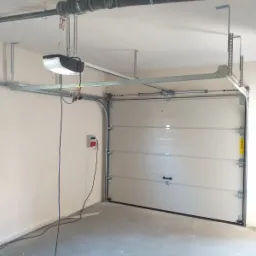 Montaż segmentowej bramy garażowej z automatycznym napędem, widoczne prowadnice, silnik z podwieszeniem, instalacja elektryczna, surowa posadzka garażu.