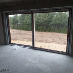 Duże przesuwne okno PCV z brązową ramą, widok na ogr&oacute;d z tujami, surowy stan wykończenia wnętrza.