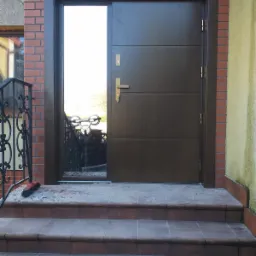 Brązowe drzwi wejściowe z bocznym przeszkleniem, widoczne przyssawki do szkła, ceglana elewacja i schody z płytek, ślady montażu.