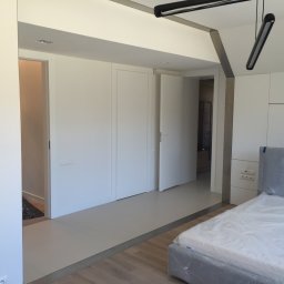 Jakub bbb - Nowoczesne wnętrze sypialni z białymi ścianami, zabudową meblową i częściowo ułożoną podłogą, łóżko zabezpieczone folią ochronną.