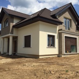 Jakub bbb - Dwukondygnacyjny dom z beżową elewacją i brązowym dachem, widoczny z perspektywy gruntu z odsłoniętą ziemią wokół budynku.