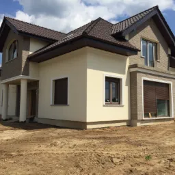 Dwukondygnacyjny dom z beżową elewacją i brązowym dachem, widoczny z perspektywy gruntu z odsłoniętą ziemią wokół budynku.