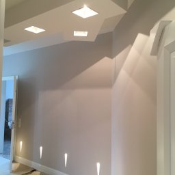 Jakub bbb - Pomieszczenie po malowaniu ścian na delikatny odcień szarości, z dekoracyjnym oświetleniem punktowym przy podłodze i zabudową sufitową z wpuszczonymi lampami.