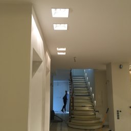 Jakub bbb - Wnętrze w trakcie odświeżania: widok na schody zabezpieczone taśmą malarską, z widocznym wałkiem do malowania i osobą w tle, oświetlone wpuszczanymi lampami sufitowymi.