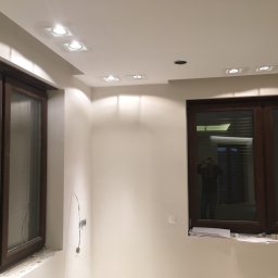 Jakub bbb - Wnętrze pokoju w trakcie prac wykończeniowych: widoczne nowe okna z brązowymi ramami, białe ściany, zamontowane punktowe oświetlenie sufitowe, niepodłączone przewody elektryczne i żarówka...