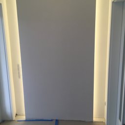 Jakub bbb - Świeżo pomalowana, szara ściana z wbudowanym oświetleniem po bokach, zabezpieczona folią malarską na podłodze.