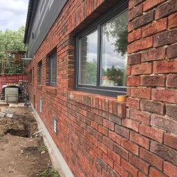 Jakub bbb - Ściana budynku z czerwonej cegły z szarymi oknami, częściowo wykończona, widoczny wykop w ziemi i rusztowanie w tle.