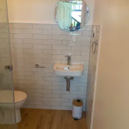 Mała łazienka z beżowymi płytkami imitującymi cegłę, drewnopodobną podłogą, okrągłym lustrem z mozaiką, białą umywalką i toaletą za szklaną kabiną prysznicową.