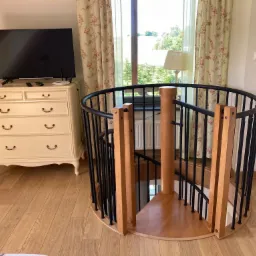 Wnętrze domu z krętymi schodami z drewnianymi stopniami i czarnymi metalowymi barierkami, obok komoda z telewizorem i figurką słonia, okno z kwiecistymi zasłonami, parkiet na podłodze.
