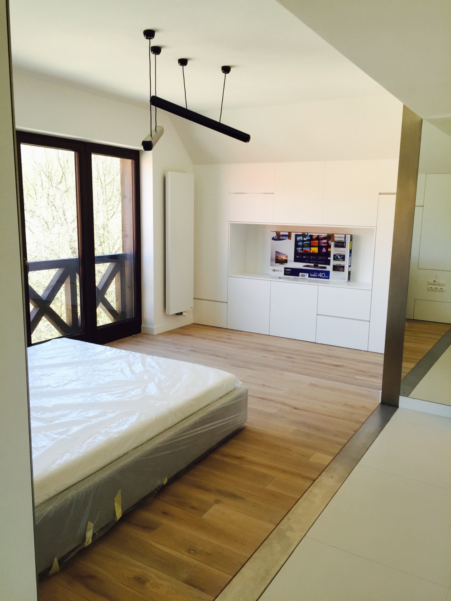 Jasne wnętrze sypialni po remoncie: drewniana podłoga, białe meble w zabudowie z miejscem na telewizor, łóżko zabezpieczone folią, nowoczesne oświetlenie, widok na zewnątrz przez drewniane okno...