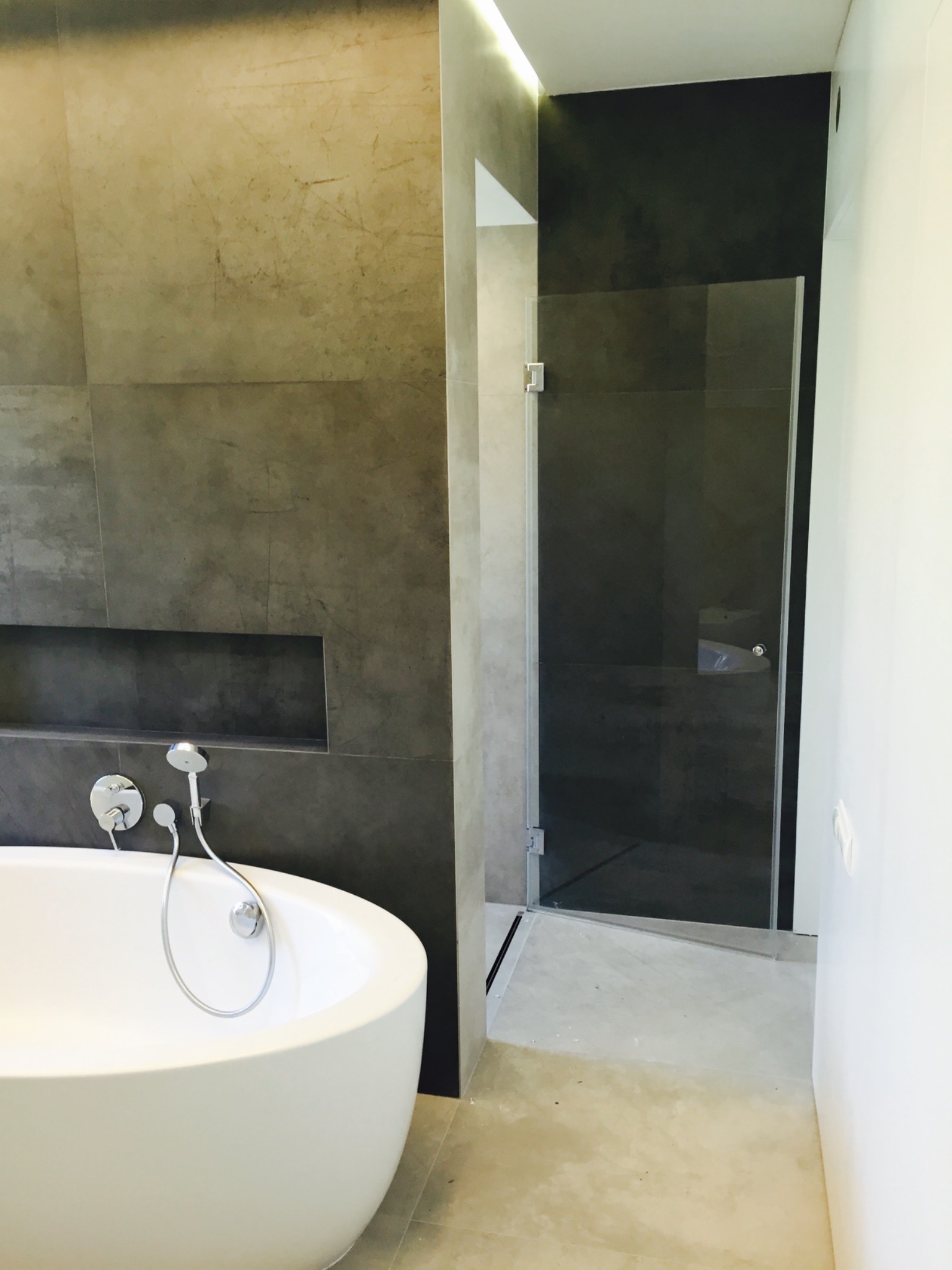 Nowoczesna łazienka z wanną wolnostojącą, szarą płytką imitującą beton i kabiną prysznicową ze szklanymi drzwiami.