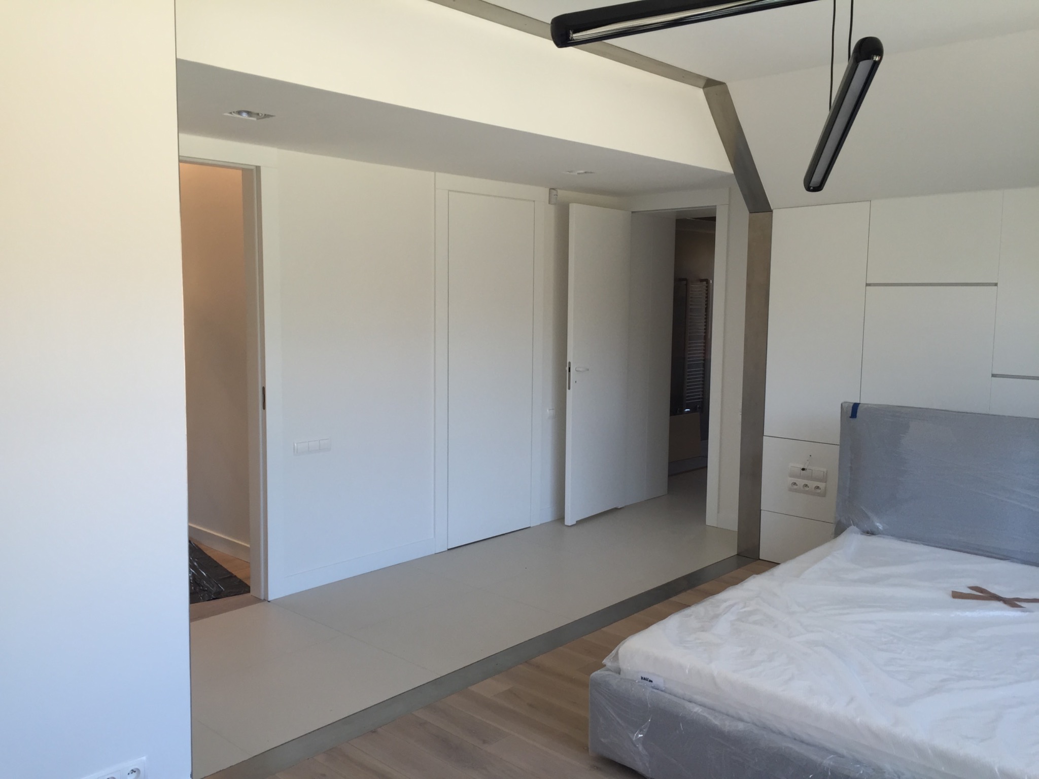 Nowoczesne wnętrze sypialni z białymi ścianami, zabudową meblową i częściowo ułożoną podłogą, łóżko zabezpieczone folią ochronną.