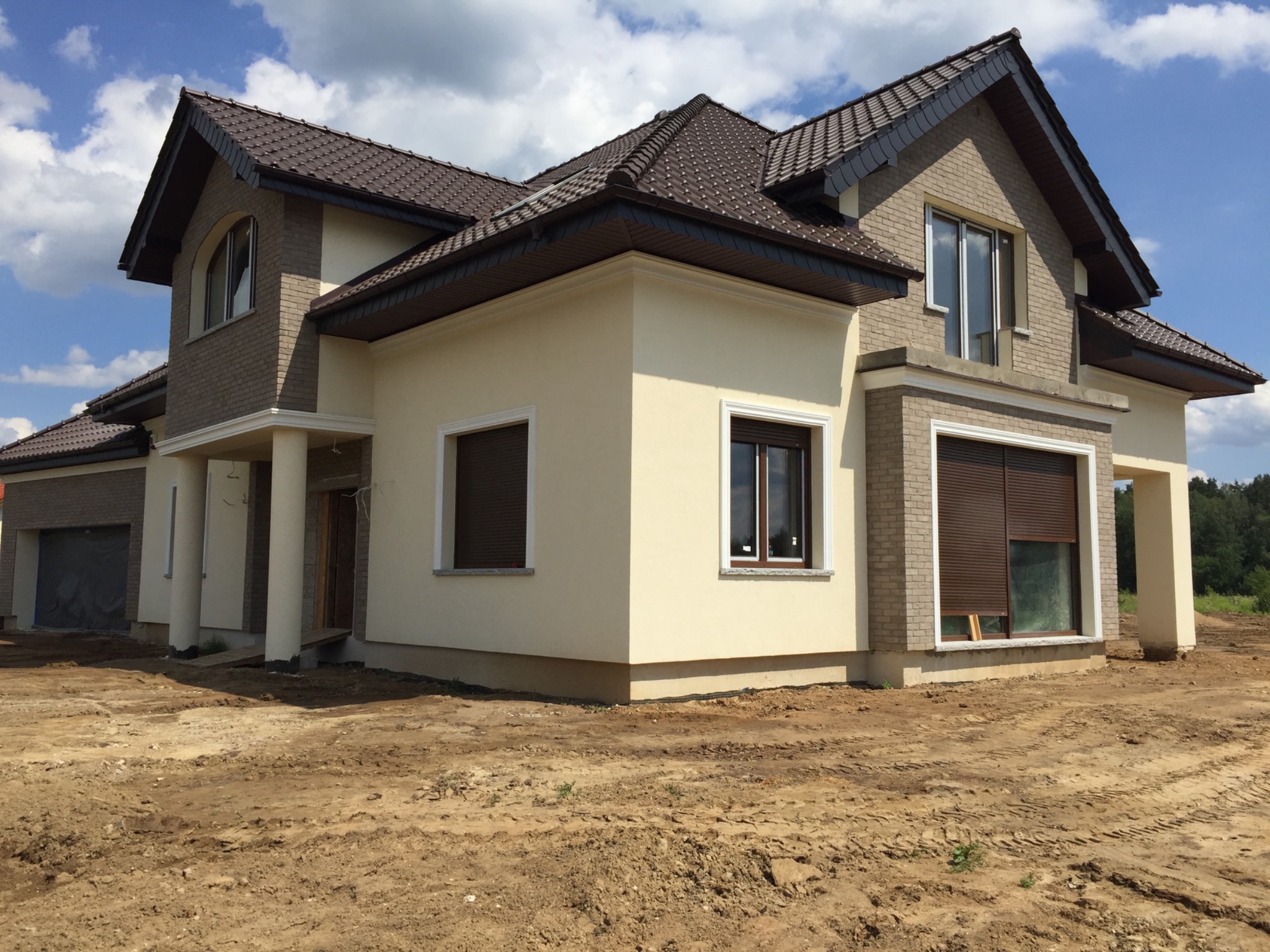 Dwukondygnacyjny dom z beżową elewacją i brązowym dachem, widoczny z perspektywy gruntu z odsłoniętą ziemią wokół budynku.