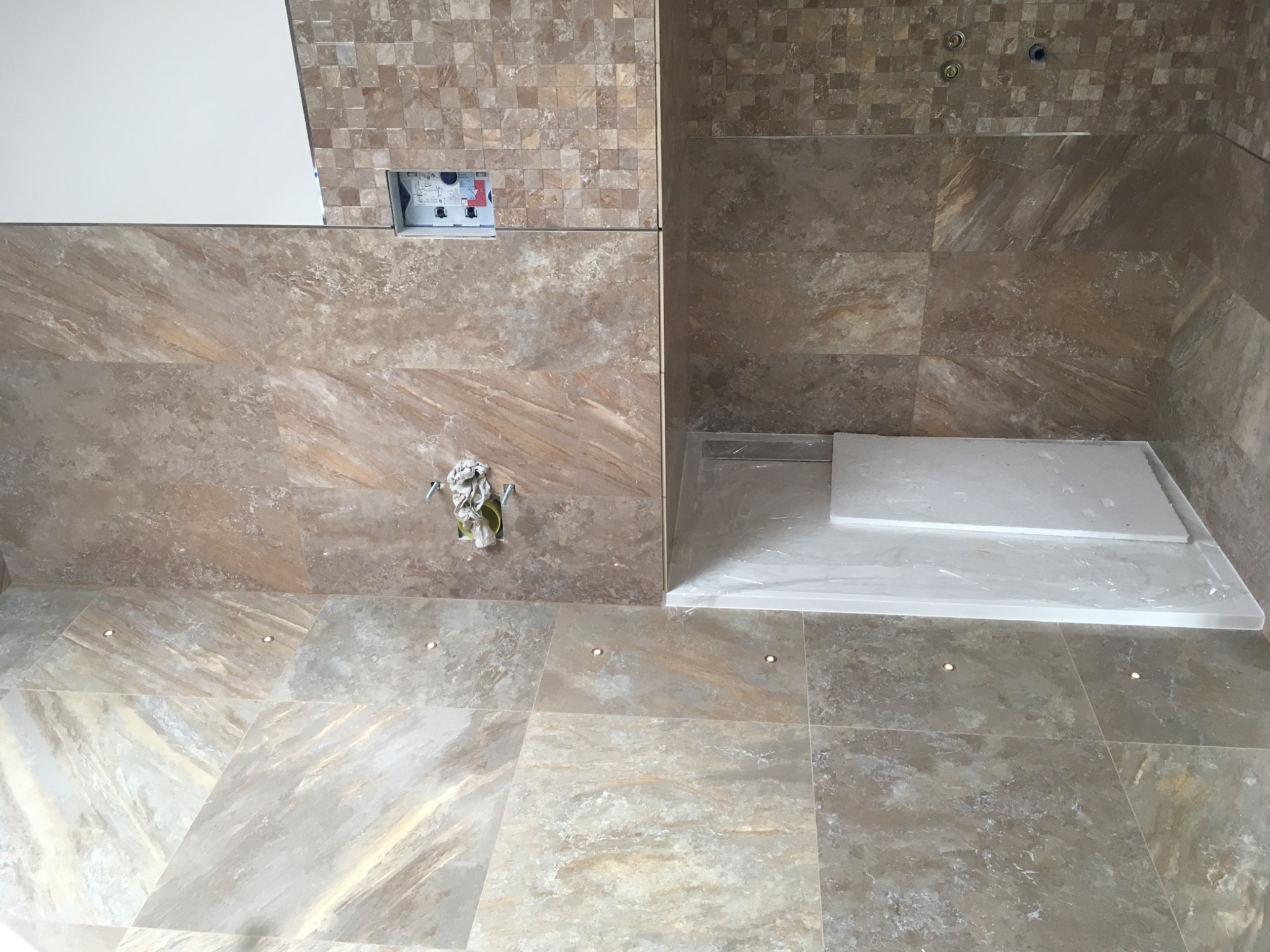 Łazienka w trakcie wykańczania z beżowymi płytkami imitującymi marmur na ścianach i podłodze, widoczny brodzik z odpływem liniowym i wystające przewody instalacji elektrycznej