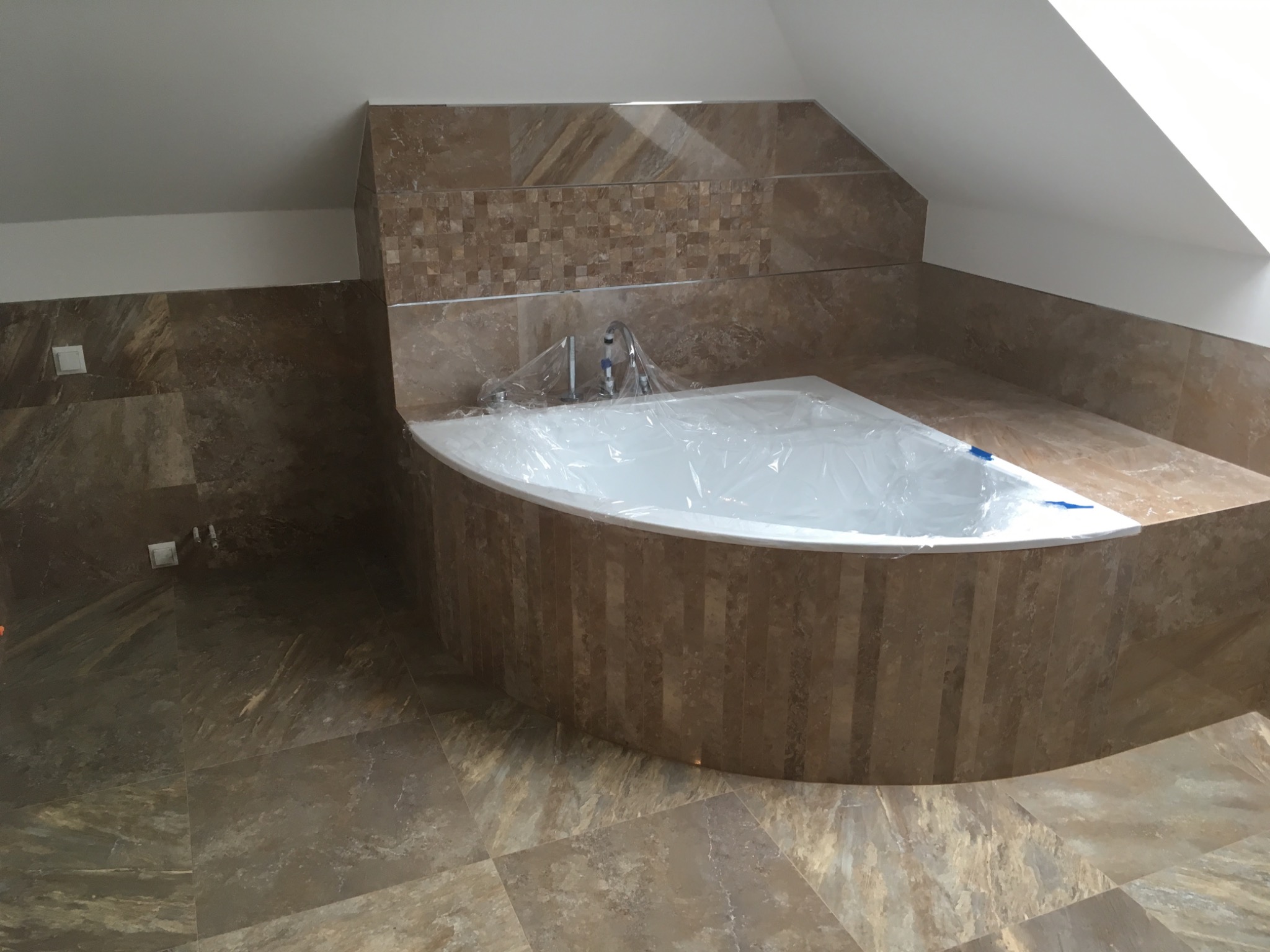 Narożna wanna obudowana pionowymi płytkami w odcieniach brązu, zabezpieczona folią ochronną, z baterią umywalkową, w łazience na poddaszu wykończonej płytkami imitującymi kamień.