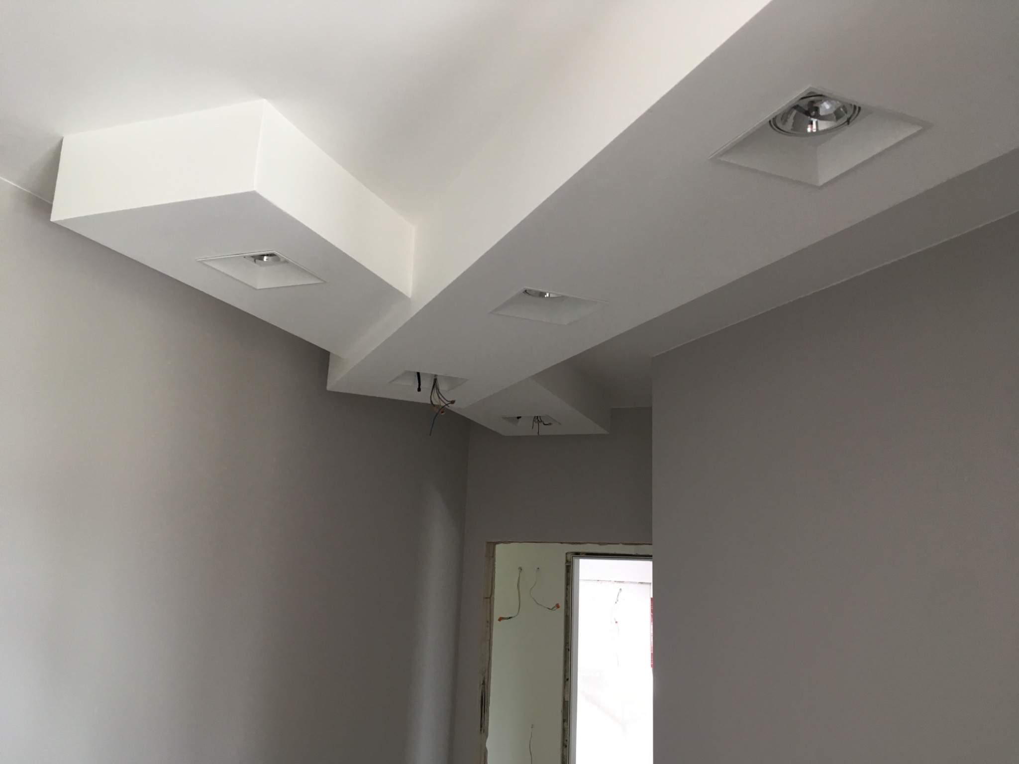 Szare ściany świeżo pomalowanego pokoju z białym sufitem podwieszanym, w którym zamontowano kwadratowe reflektory punktowe. Widoczne wystające kable z dwóch otworów na reflektory.