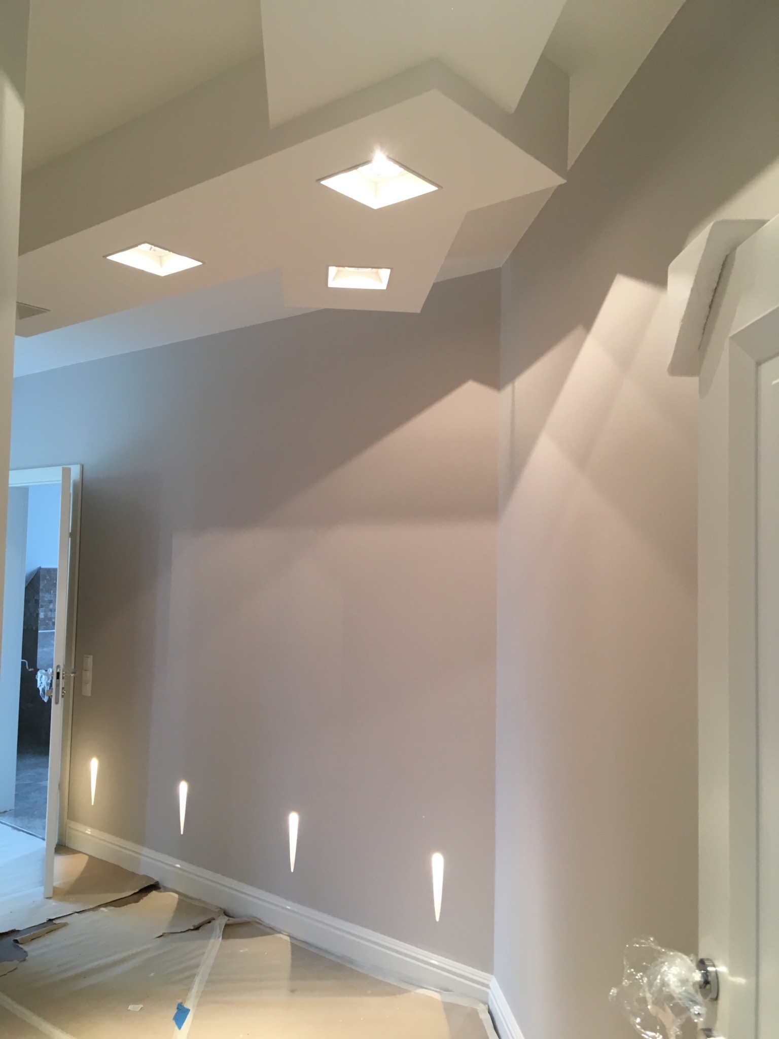 Pomieszczenie po malowaniu ścian na delikatny odcień szarości, z dekoracyjnym oświetleniem punktowym przy podłodze i zabudową sufitową z wpuszczonymi lampami.