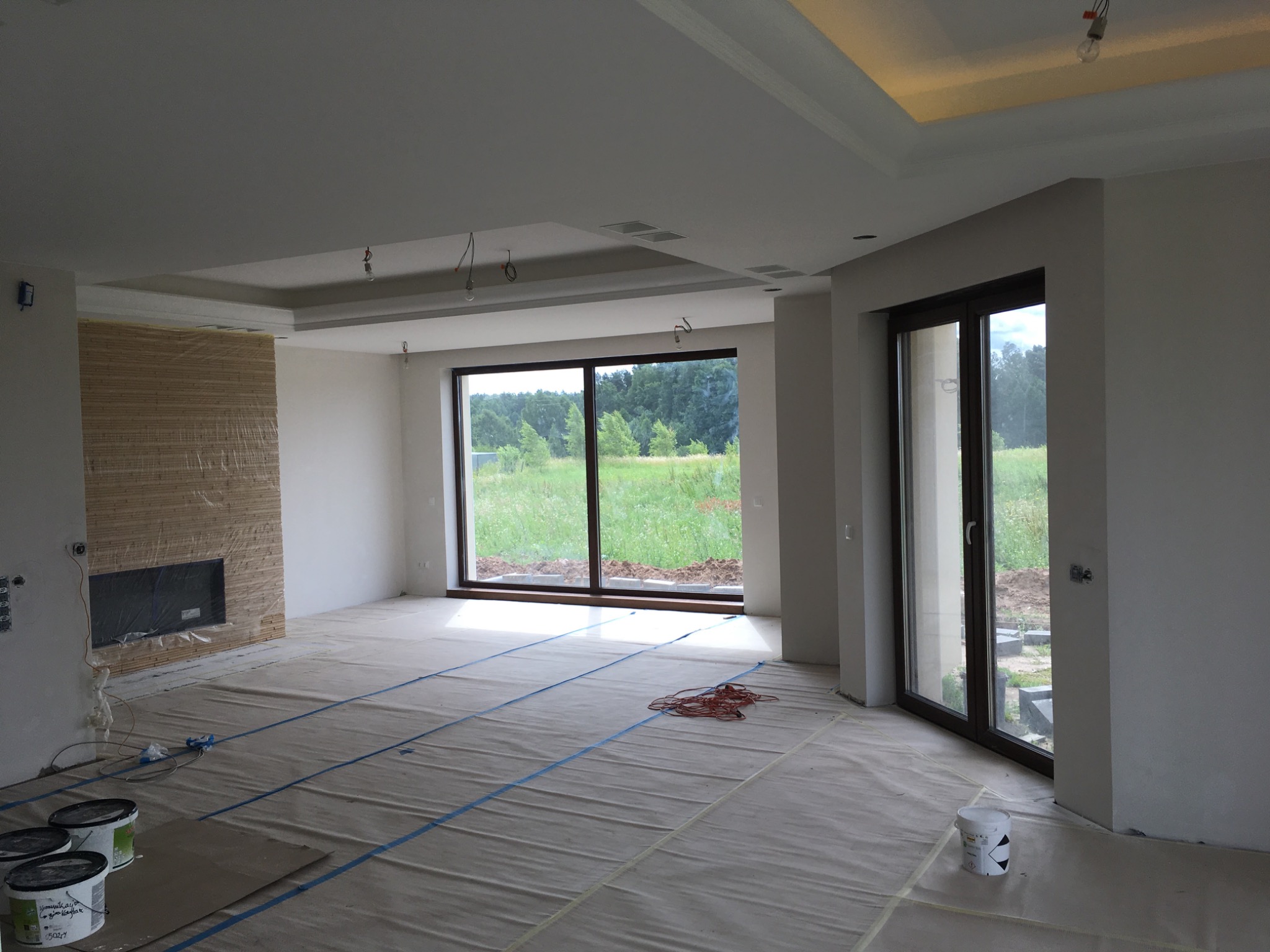 Surowe wnętrze domu w trakcie wykończenia: zabezpieczona folią podłoga, ściana z imitacją drewna, duże okna z widokiem na zieleń, widoczne puszki z farbą.