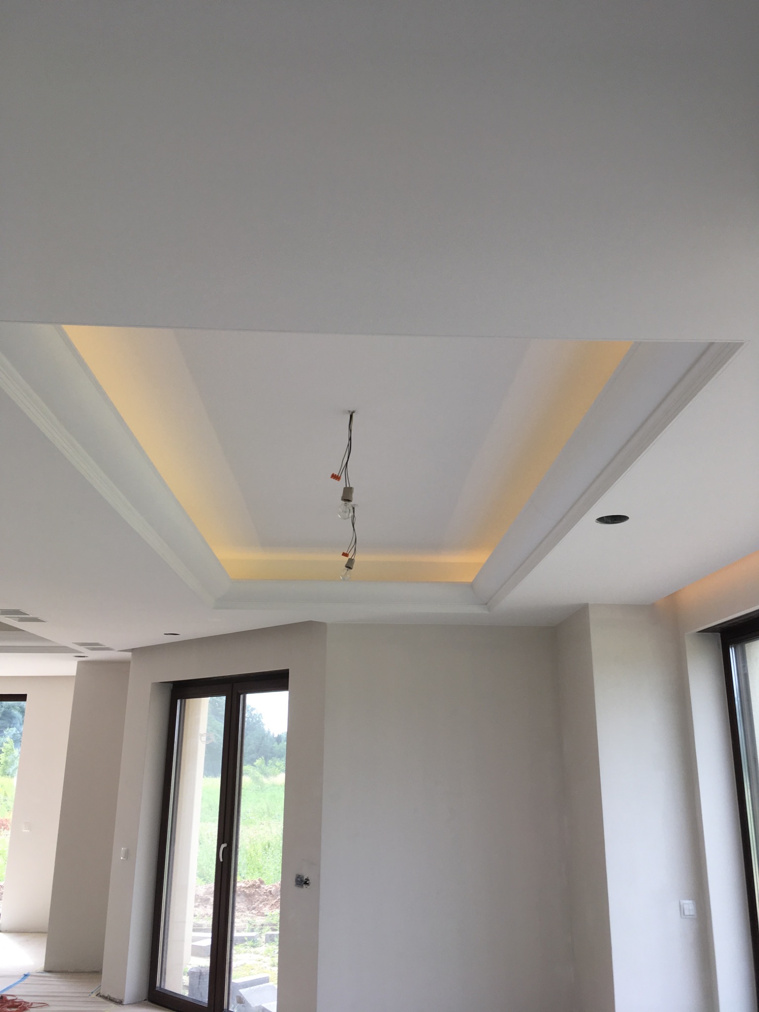 Nowoczesny sufit podwieszany z oświetleniem LED w nowym, jasnym pomieszczeniu z dużymi oknami balkonowymi, widoczne kable wystające z sufitu w miejscu planowanego oświetlenia.