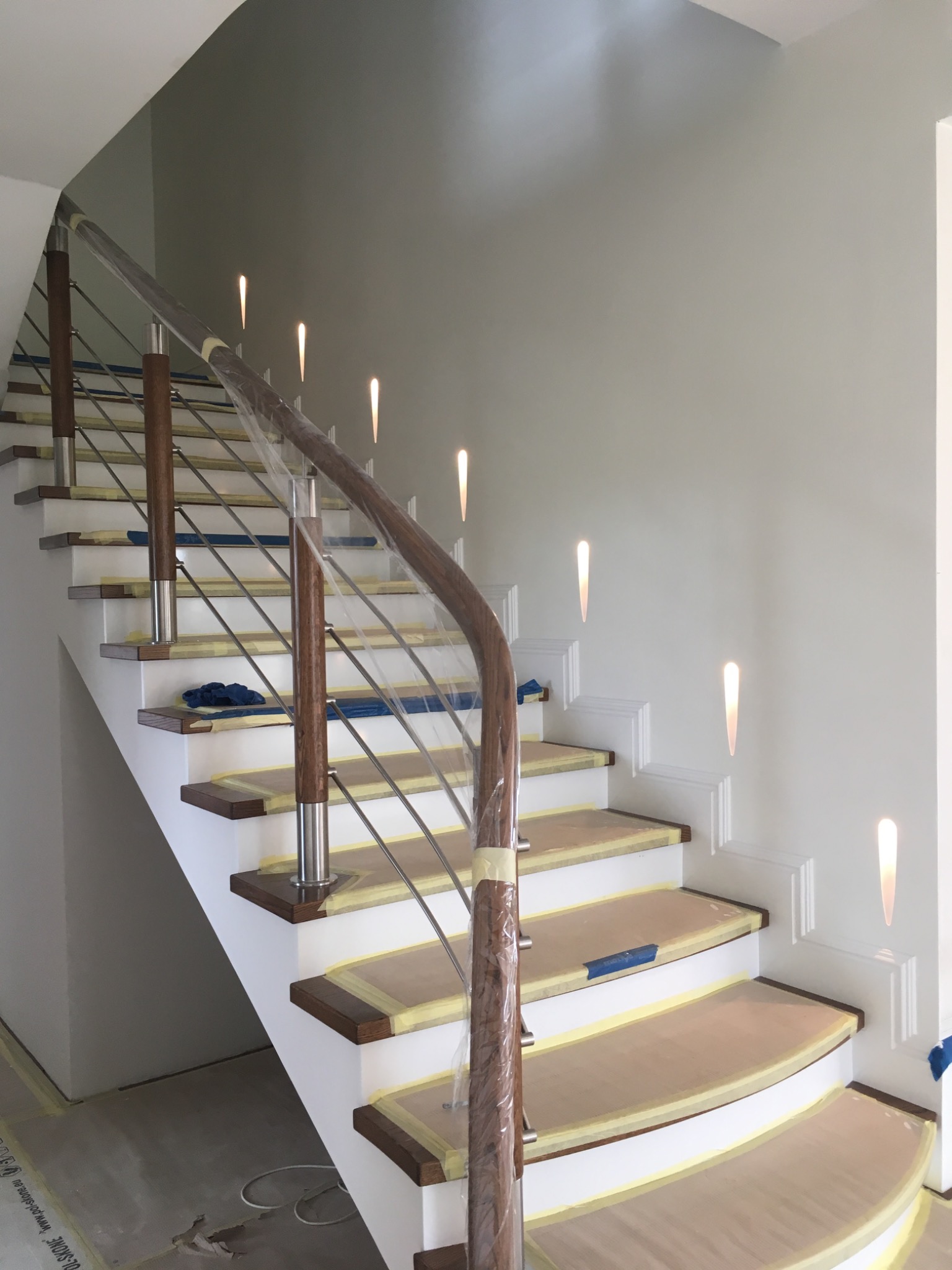 Schody z drewnianymi stopniami i metalową balustradą w trakcie zabezpieczania folią malarską w Gdańsku, widoczne oświetlenie ledowe w ścianie.