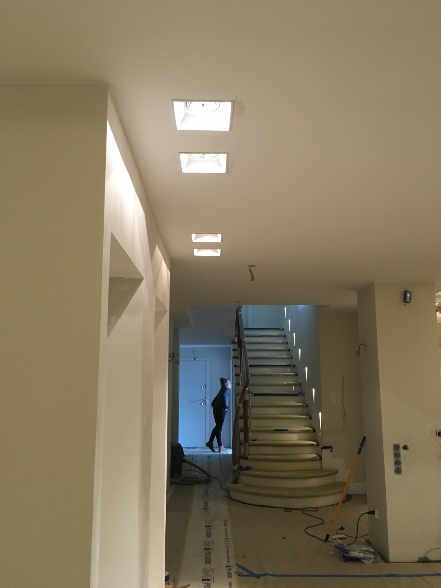 Wnętrze w trakcie odświeżania: widok na schody zabezpieczone taśmą malarską, z widocznym wałkiem do malowania i osobą w tle, oświetlone wpuszczanymi lampami sufitowymi.