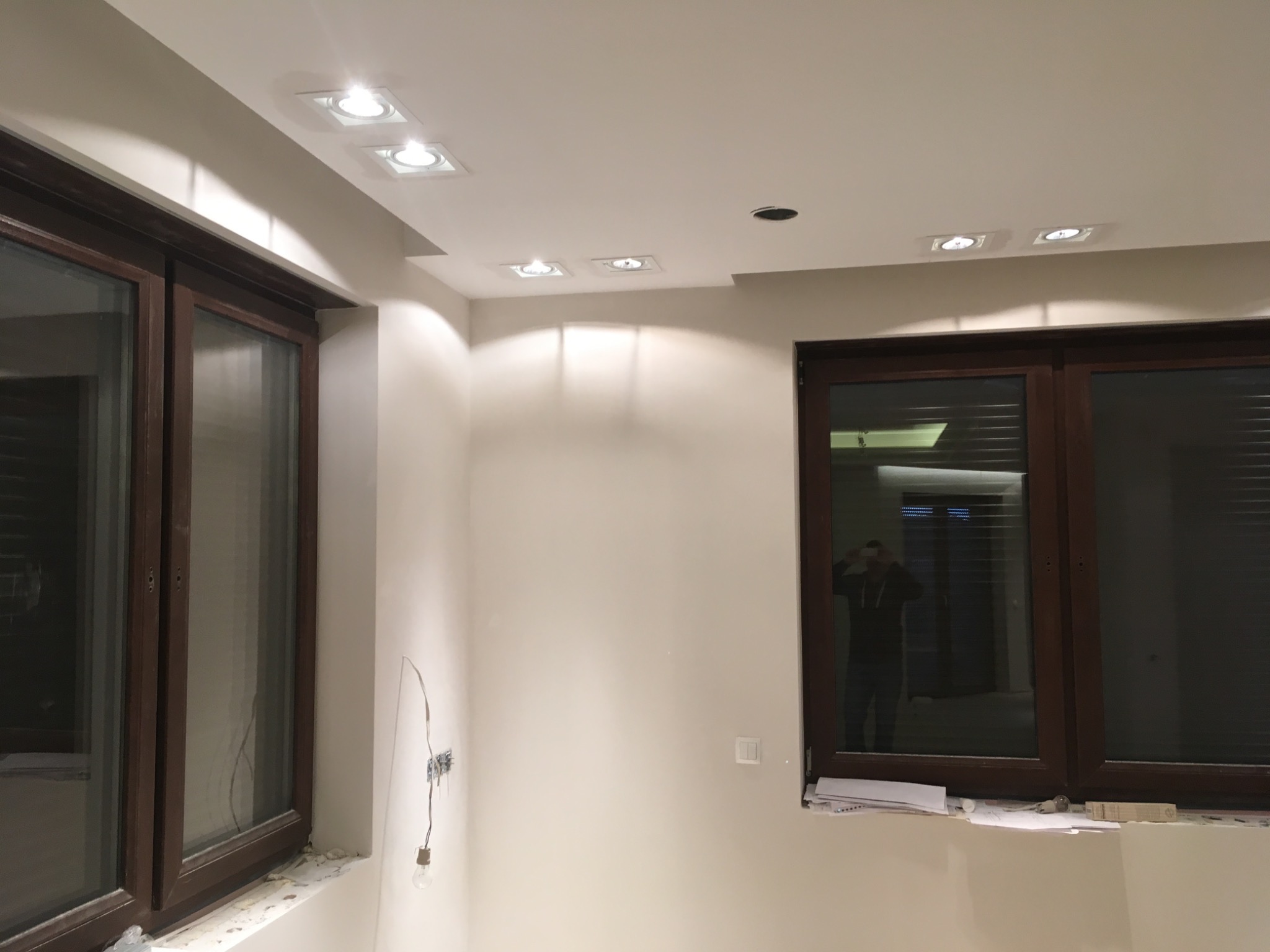 Wnętrze pokoju w trakcie prac wykończeniowych: widoczne nowe okna z brązowymi ramami, białe ściany, zamontowane punktowe oświetlenie sufitowe, niepodłączone przewody elektryczne i żarówka...