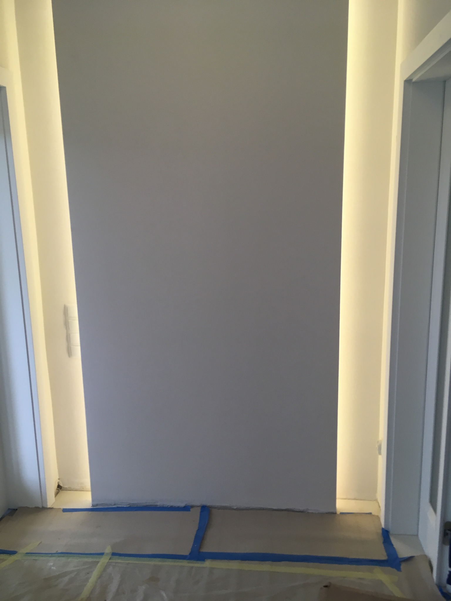 Świeżo pomalowana, szara ściana z wbudowanym oświetleniem po bokach, zabezpieczona folią malarską na podłodze.