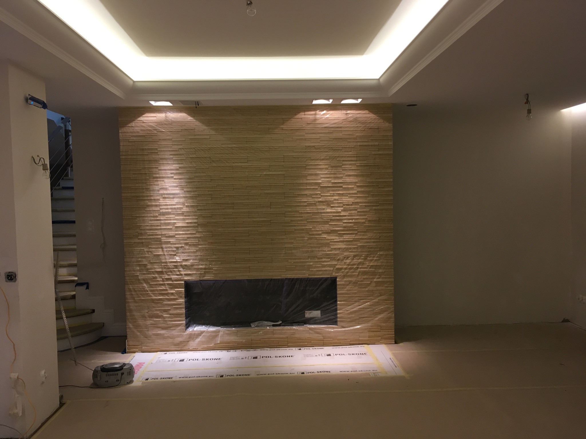 Ściana z dekoracyjnej okładziny imitującej kamień, z wnęką na kominek, zabezpieczona folią ochronną podczas prac wykończeniowych, widoczne przygotowane podłoże i podświetlany sufit.