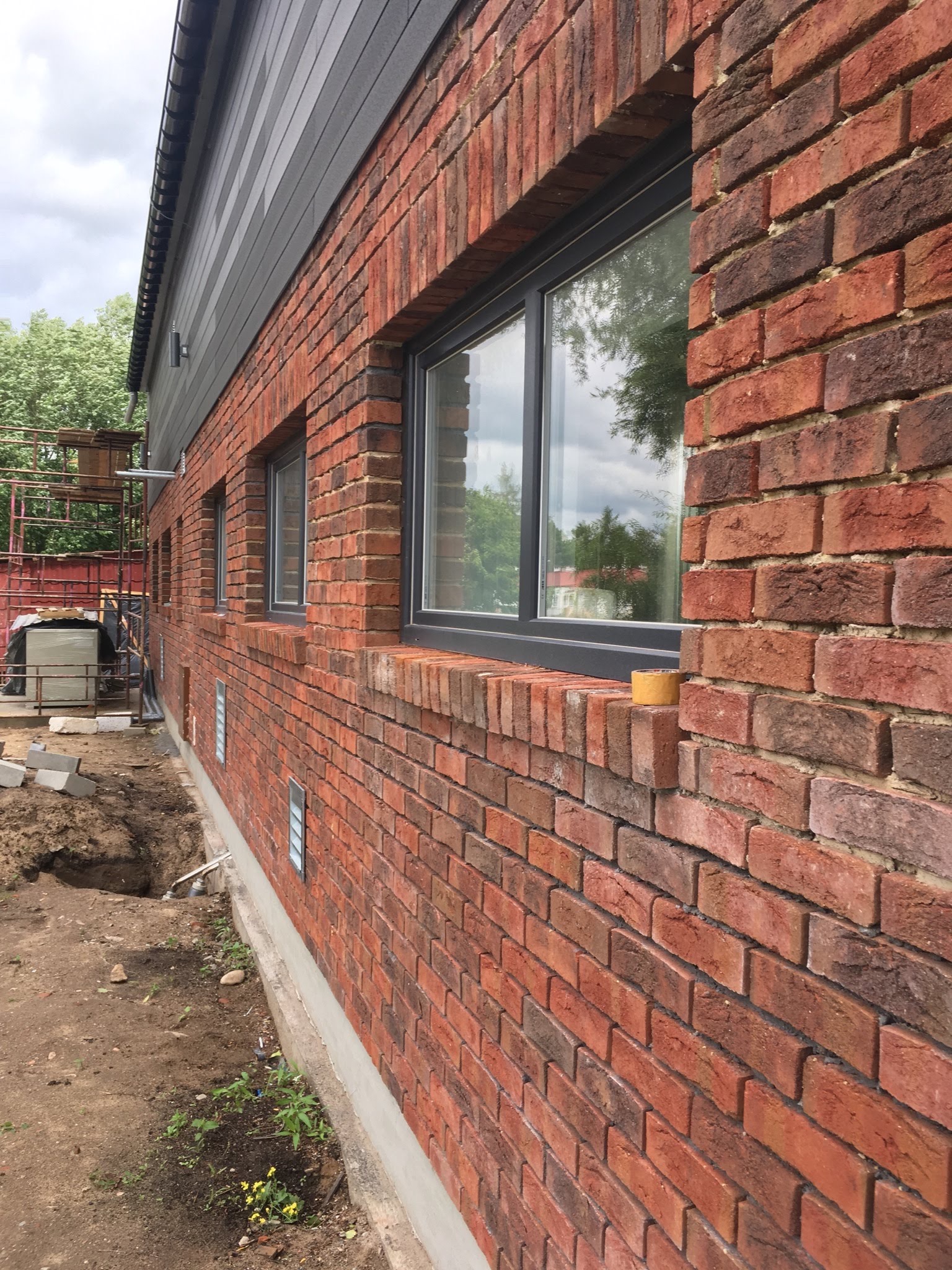 Ściana budynku z czerwonej cegły z szarymi oknami, częściowo wykończona, widoczny wykop w ziemi i rusztowanie w tle.