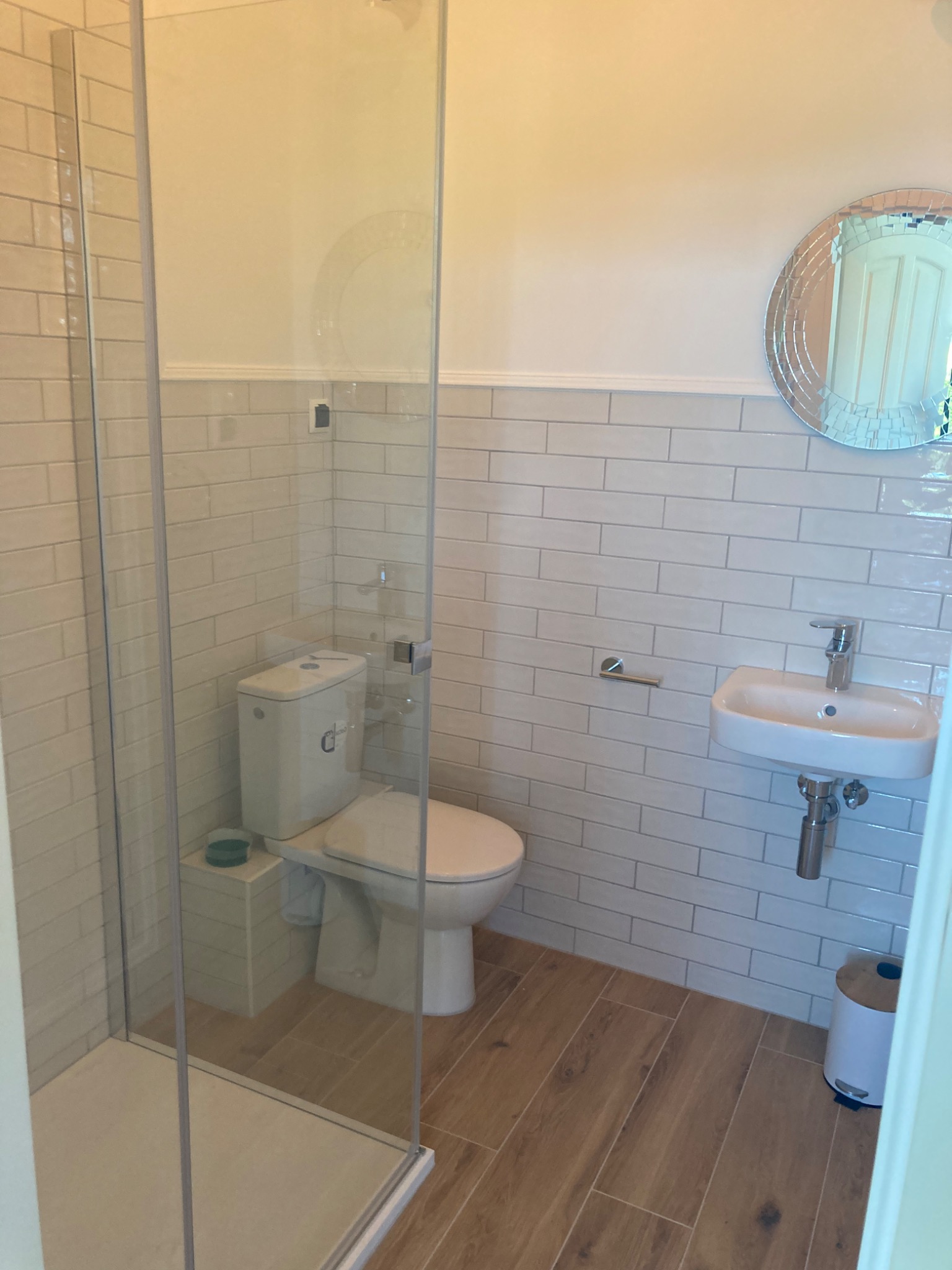 Nowoczesna łazienka z kabiną prysznicową ze szkła, podwieszaną toaletą, umywalką i lustrem w ramie z małych luster, wykończona beżowymi płytkami imitującymi cegłę i drewnopodobną podłogą.